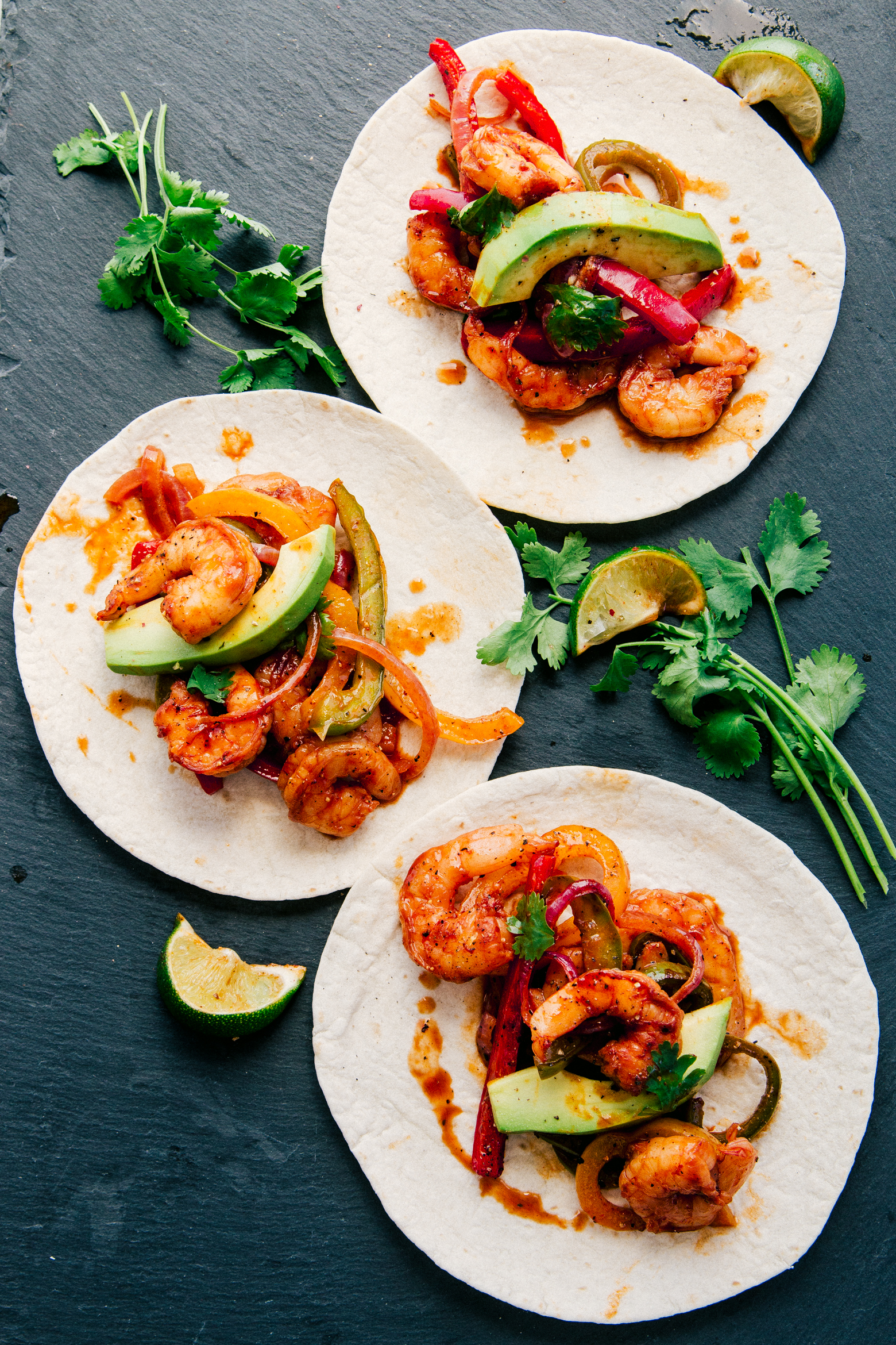 Shrimp Fajitas served on tortillas with sliced avocados and cilantro on a black background by The Food Cafe. 