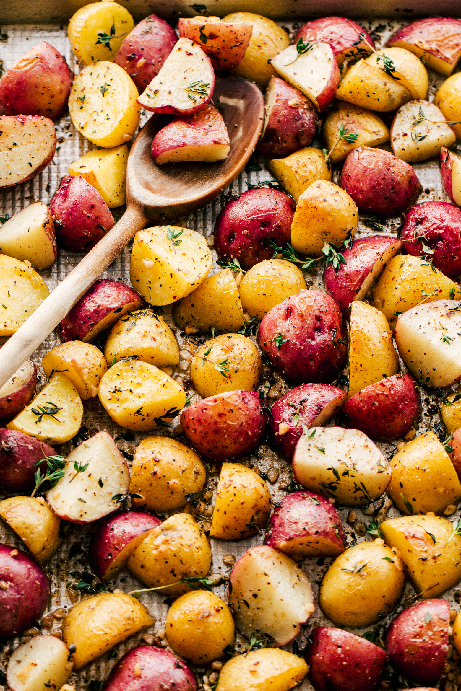 Roasted Potatoes on a sheet pan with a wooden spoon for serving topped with garlic butter and chopped rosemary by The Food Cafe