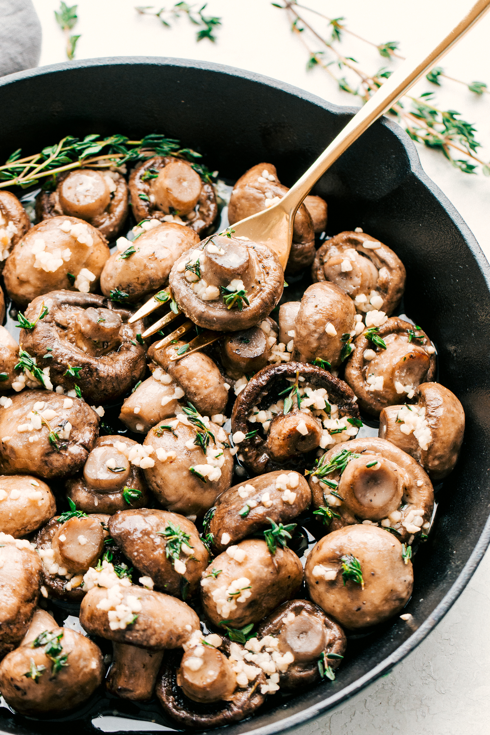 Roasted mushrooms in garlic butter in a black cast iron skillet served on a white background and a gold fork, by The Food Cafe. 