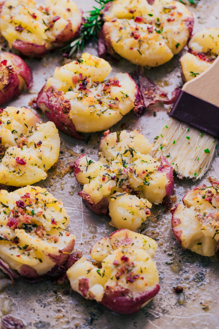 Smashed potatoes on a dark sheet pan with a brush, brushing on butter. 