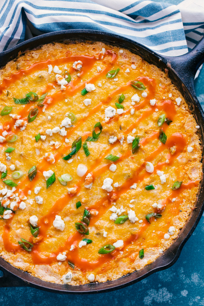 Buffalo Chicken Dip in a skillet topped with blue cheese crumbles, hot sauce and chopped green onions, perfect for your super bowl party by The Food Cafe. 
