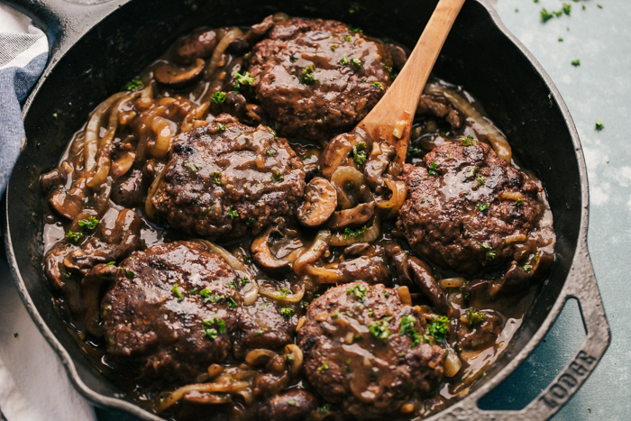 hamburger steaks in a skillet being topped with mushroom and onion gravy. 