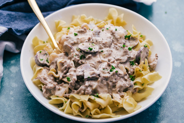 hamburger stroganoff recipe served in a white bowl with spoon on a blue background by The Food Cafe. 