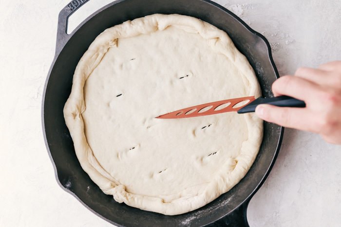 Pizza dough in a cast iron skillet with several 1 inch slits cut with a knife into it to allow the dough to cook to make meatball pizza by The Food Cafe. 