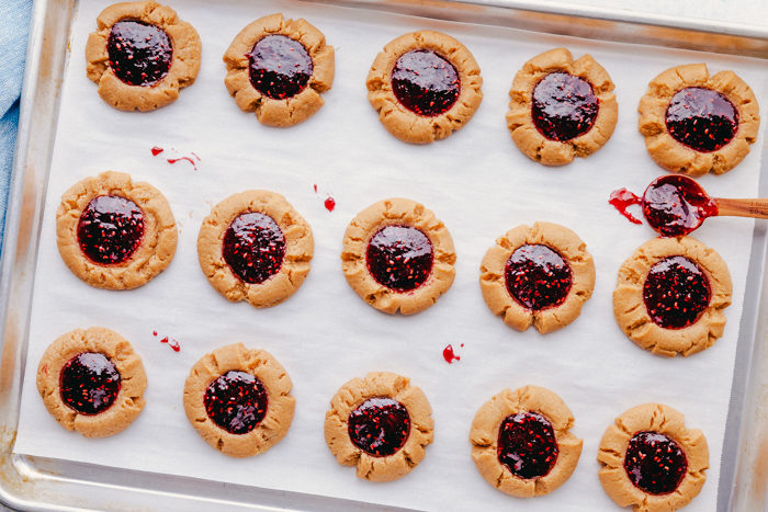 Peanut butter thumbprint cookies layered on a silver cookie sheet with parchment paper topped with raspberry jam, by The Food Cafe