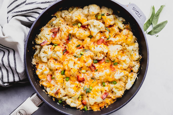 Cheesy cauliflower casserole in a skillet on a white background garnished with diced bacon and green onions, done in 20 minutes, by The Food Cafe. 