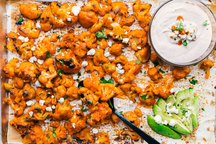 Buffalo cauliflower bites made on a rimmed sheet pan garnished with crumbled blue cheese, cilantro and avocados, served with a spicy ranch dip in a clear bowl, by The Food Cafe. 