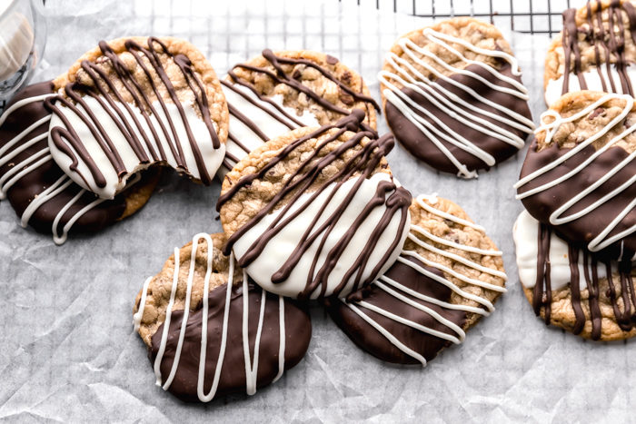 Chocolate dipped cookies dipped in dark chocolate and white chocolate with additional chocolate drizzled over the top on a piece of white parchment paper sitting on a black cooling rack with a tall glass of milk along side, by the Food Cafe. 