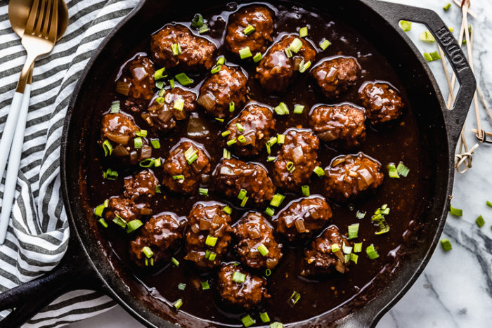 Baked meatballs in a black skillet placed in Guinness sauce on white background garnished with diced green onions sitting on a grey and white stripped linen napkin served with toothpicks and a gold fork and spoon, by The Food Cafe.