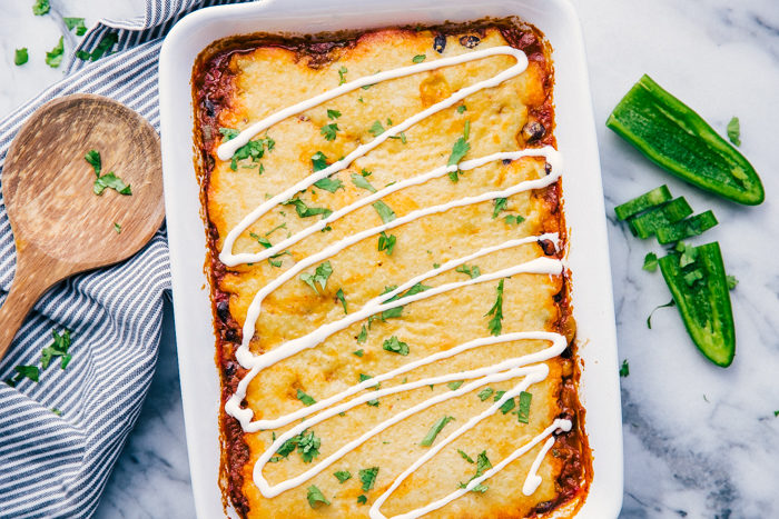 Mexican cornbread casserole in a white casserole dish on a white background with a wooden serving spoon on the left and diced jalapenos on the right, topped with cilantro and sour cream, by The Food Cafe