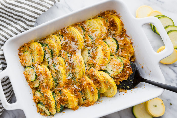 squash and zucchini casserole in a rectangular white casserole dish with a black serving spoon laid on a light background with fresh sliced zucchini garnished around the dish, by The Food Cafe. 