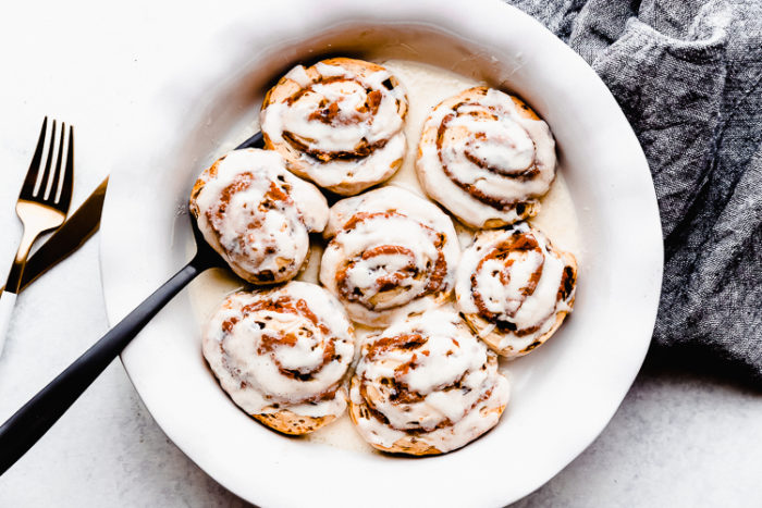 Pumpkin cinnamon rolls in a white serving dish topped with cream cheese icing on a white background and a black serving spatula, quick to make by The Food Cafe.