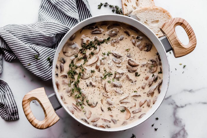 A white background with a stock pot filled with homemade cream of mushroom soup sitting on a blue and white napkin with fresh thyme sprinkled around the pot, by The Food Cafe. 